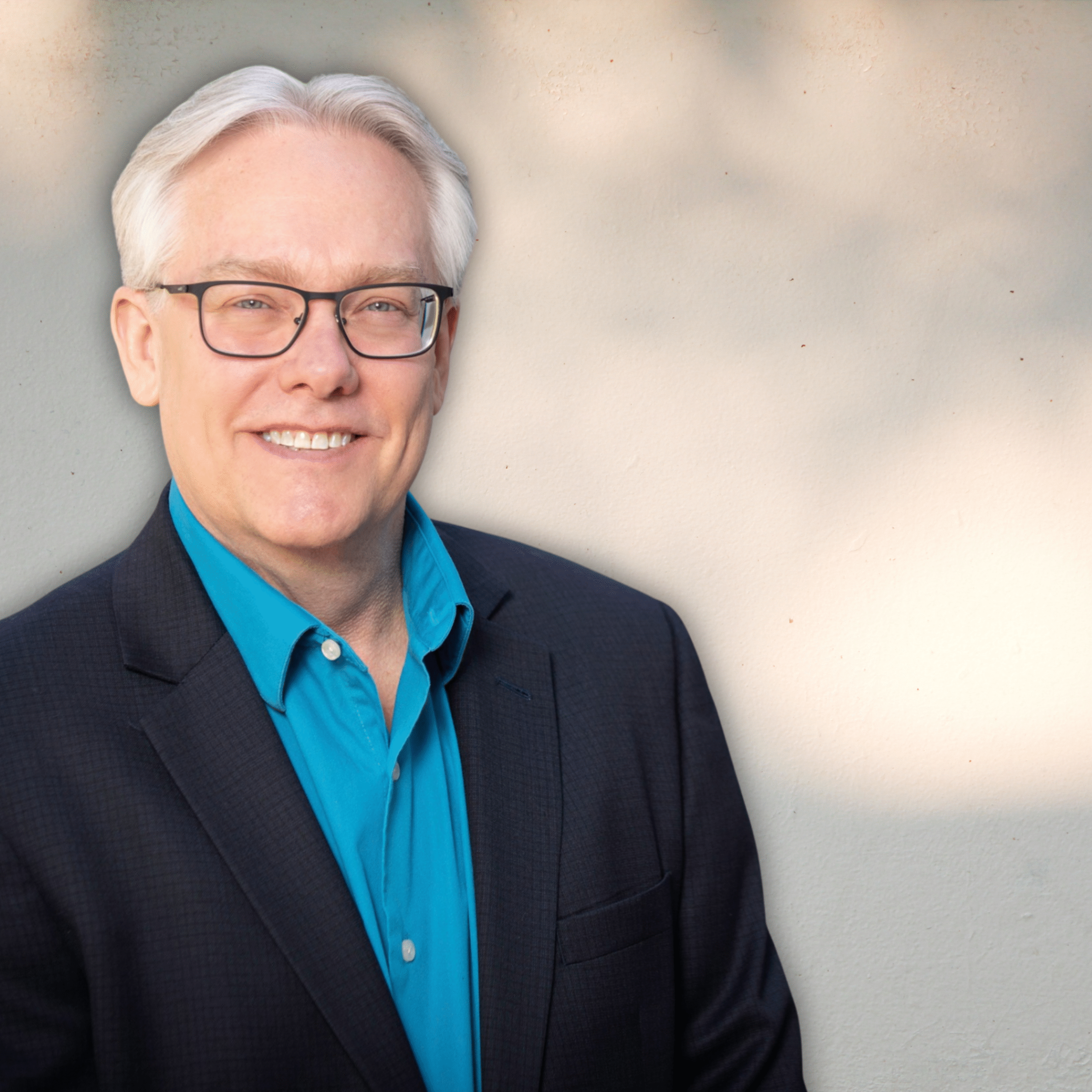 A smiling man with white hair and glasses wearing a navy blazer and blue shirt, standing against a neutral background.