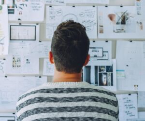 Person standing in front of a wall of scattered plans and notes, representing strategy misalignment, information overload, and lack of clear direction.