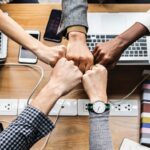 Diverse hands come together in a fist bump over a wooden desk with laptops and phones, signaling teamwork.