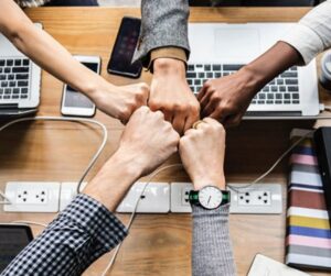 Diverse hands come together in a fist bump over a wooden desk with laptops and phones, signaling teamwork.